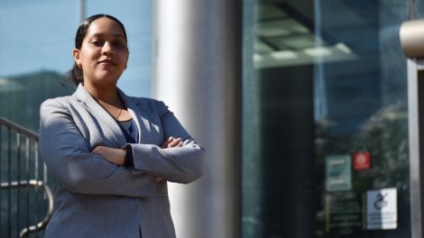 A law student interning at the Queens District Attorney's Office stands outside the office building with her arms crossed, wearing a gray blazer over business attire. She faces the camera with a confident expression as the glass façade of the courthouse reflects the sky behind her.