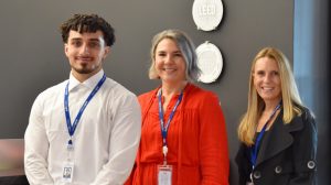 Three National Grid employees wearing ID badges smile together in an office. The man on the left wears a white shirt, the woman in the center wears a red dress, and the woman on the right wears a black blazer.